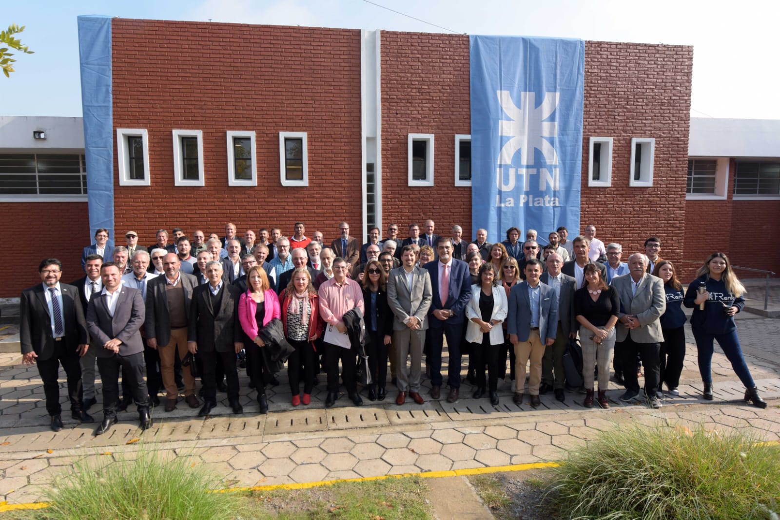 La imagen describe una foto grupal del Consejo Superior de la UTN reunido en la Facultad Regional de La Plata, detrás la fachada del edificio y una bandera con el logo de la UTN.